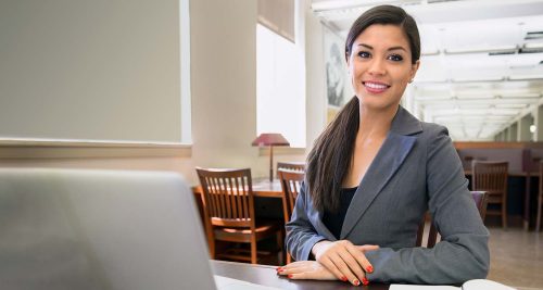 Smiling, successful looking woman in gray blazer sitting at desk in front of laptop.